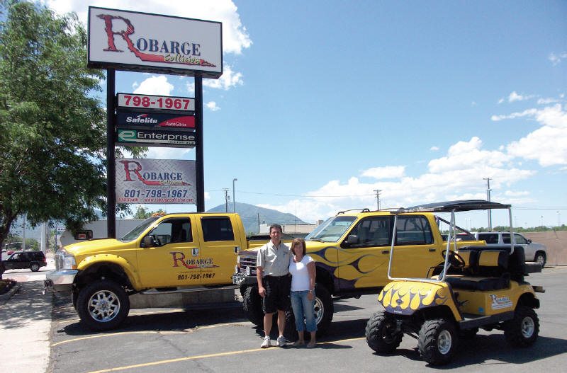 2 people standing in front of company sign and yellow truck, golf cart & 4 wheeler